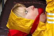 Pia tied and gagged in bed in a yellow rainsuit and a red gag