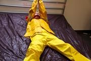 Pia tied and gagged in bed in a yellow rainsuit and a red gag