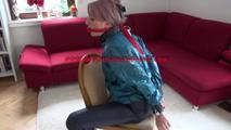Mara tied and gagged on a chair with a bar wearing a sexy blue shiny nylon rain pants and a green rain jacket (Video)