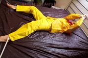 Pia tied and gagged in bed in a yellow rainsuit and a red gag