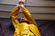 Pia tied and gagged in bed in a yellow rainsuit and a red gag