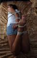 Barefoot Christy in white top and jeans shorts, hogtied with her elbows tied together, is tickled and teased by camera man