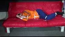 Lucy wearing a blue rain pants and an orange rain jacket being tied and gagged with belts on a sofa (Video)