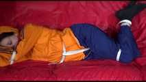 Lucy wearing a blue rain pants and an orange rain jacket being tied and gagged with belts on a sofa (Video)