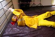 Pia tied and gagged in bed in a yellow rainsuit and a red gag