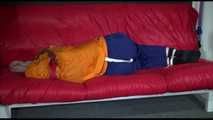 Lucy wearing a blue rain pants and an orange rain jacket being tied and gagged with belts on a sofa (Video)