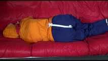 Lucy wearing a blue rain pants and an orange rain jacket being tied and gagged with belts on a sofa (Video)