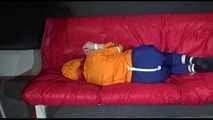 Lucy wearing a blue rain pants and an orange rain jacket being tied and gagged with belts on a sofa (Video)