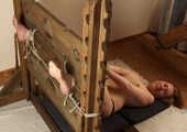 Topless barefoot Florence, lying on the floor with her hands tied in front of her and with her feet locked in stocks, is tickled by a man