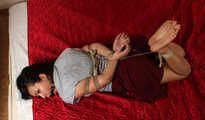 Ballgagged barefoot Lois in grey t-shirt and red skirt, hogtied with hemp rope, rolls and wiggles on the mattress