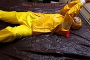 Pia tied and gagged in bed in a yellow rainsuit and a red gag
