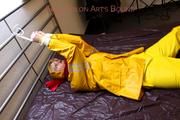 Pia tied and gagged in bed in a yellow rainsuit and a red gag