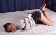 Barefoot Lisa in white shirt and jeans shorts, hogtied with hemp rope, wiggles and rolls on the bed
