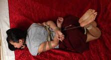 Ballgagged barefoot Lois in grey t-shirt and red skirt, hogtied with hemp rope, rolls and wiggles on the mattress