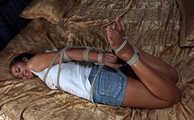 Barefoot Christy in white top and jeans shorts, hogtied with her elbows tied together, is tickled and teased by camera man