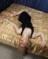 Barefoot blonde Alina in black dress, hog-cuffed with leather cuffs, wiggles on the bed