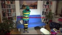 Mara tied, gagged and hooded with tape on a blue sofa wearing an blue/green rain combination (Video)