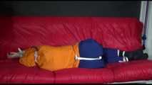 Lucy wearing a blue rain pants and an orange rain jacket being tied and gagged with belts on a sofa (Video)
