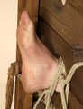 Topless barefoot Florence, lying on the floor with her hands tied in front of her and with her feet locked in stocks, is tickled by a man