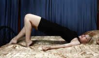 Barefoot blonde Alina in black dress, hog-cuffed with leather cuffs, wiggles on the bed