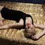Barefoot blonde Alina in black dress, hog-cuffed with leather cuffs, wiggles on the bed