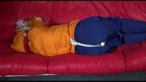 Lucy wearing a blue rain pants and an orange rain jacket being tied and gagged with belts on a sofa (Video)