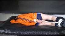 Lucy tied and gagged on a bed wearing a blue shiny nylon shorts and an orange shiny nylon rain jacket (Video)