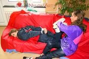 Leonie tied and gagged by Stella on a sofa both wearing sexy shiny nylon rainwear (Pics)
