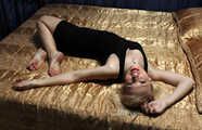 Barefoot blonde Alina in black dress, hog-cuffed with leather cuffs, wiggles on the bed