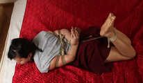 Ballgagged barefoot Lois in grey t-shirt and red skirt, hogtied with hemp rope, rolls and wiggles on the mattress
