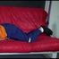 Lucy wearing a blue rain pants and an orange rain jacket being tied and gagged with belts on a sofa (Video)