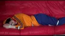 Lucy wearing a blue rain pants and an orange rain jacket being tied and gagged with belts on a sofa (Video)
