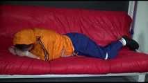 Lucy wearing a blue rain pants and an orange rain jacket being tied and gagged with belts on a sofa (Video)