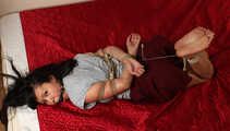 Ballgagged barefoot Lois in grey t-shirt and red skirt, hogtied with hemp rope, rolls and wiggles on the mattress