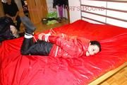 Lucy tied, gagged and hooded on a bed wearing a sexy black shiny nylon pants and a red down jacket (Pics)