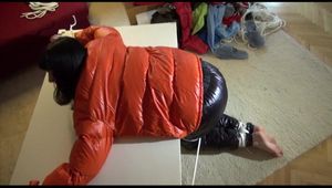 Mara tied and gagged on a table wearing a sexy black down pants and an orange down jacket (Video)