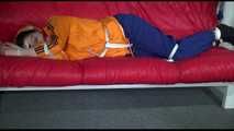 Lucy wearing a blue rain pants and an orange rain jacket being tied and gagged with belts on a sofa (Video)