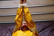 Pia tied and gagged in bed in a yellow rainsuit and a red gag
