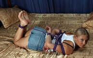 Barefoot Nancy in white tank-top and jeans shorts, hogtied with violet hemp rope, wiggles on the bed